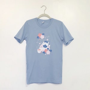 Peut inclure: Un t-shirt bleu clair avec un motif de deux chats de dessin animé buvant du thé et entourés de fleurs roses.