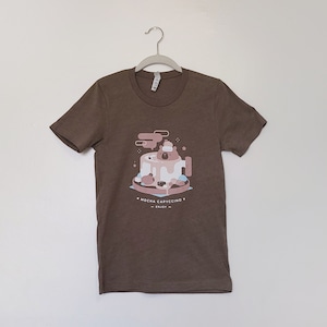 Peut inclure: Un t-shirt marron avec une illustration de dessin animé d'un ours dans une tasse de café. Le texte "MOCHA CAPPUCCINO" et "ENJOY" est imprimé sur le t-shirt.