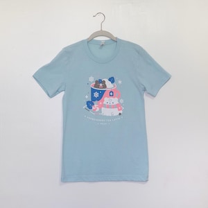 Peut inclure: T-shirt bleu clair avec un motif d'ours et d'ours polaire dégustant un latte au thé à la menthe poivrée. Le motif comprend une tasse bleue, des flocons de neige et le texte "A Peppermint Tea Latte".