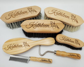 May include: A collection of personalized wooden horse grooming tools. The tools include brushes, a comb, and a hoof pick, all engraved with the name "Kathleen" and a horse and rider silhouette. The brushes have black and gray bristles.