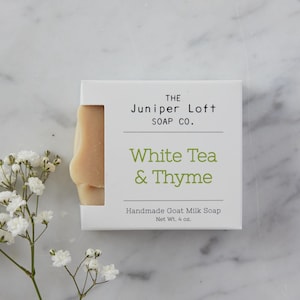 May include: A white box of handmade goat milk soap from The Juniper Loft Soap Co. The label reads "White Tea & Thyme" and "Net Wt. 4 oz." A portion of the light-colored soap is visible. White flowers are in the lower left corner.