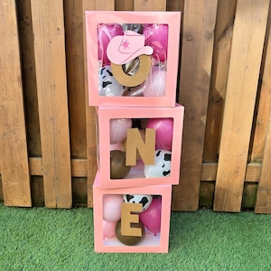 Peut inclure: Trois boîtes roses empilées épelant "ONE", chacune contenant des ballons. La boîte supérieure présente un chapeau de cow-boy rose et la lettre "O". Les ballons sont roses, marron et imprimés vache. Les boîtes sont sur l'herbe avec une clôture en bois en arrière-plan.