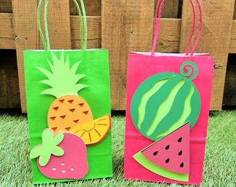 May include: Two colorful gift bags with fruit designs. One bag is green with a pineapple and strawberry, the other is pink with a watermelon. Both bags have matching colored handles and are on a grassy surface.