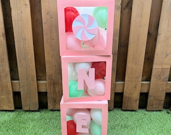 May include: Three stacked pink boxes with clear windows, filled with balloons in red, pink, and green. The top box features a swirl design, while the middle and bottom boxes display the letters "N" and "E". The boxes are arranged on green grass.