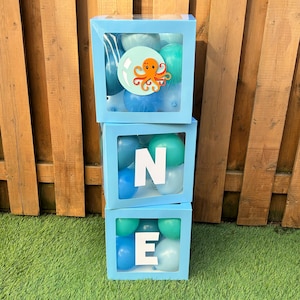 May include: Three stacked light blue transparent boxes filled with assorted blue and teal balloons. The top box features a circular cutout with an orange octopus graphic. The middle box displays a white letter N, and the bottom box displays a white letter E.