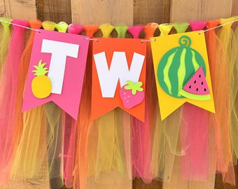 May include: A colorful banner with the word "TW" in white letters, decorated with a pineapple, strawberry, and watermelon. The banner is made of pink, orange, and yellow paper pennants and tulle, strung on a white string. The background is a wooden fence.