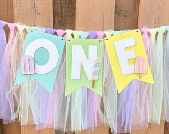 May include: A decorative banner featuring three pennants in light blue, light green, and yellow, each displaying a large white letter to spell the word ONE. The banner is adorned with small popsicle cutouts and a fringe of pastel tulle fabric.