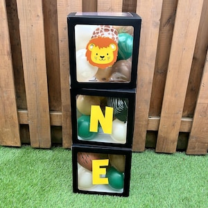 May include: Three black square boxes stacked vertically, spelling "ONE" with yellow letters. Each box contains balloons in various colours and patterns, including a lion face cutout in the top box. The boxes are set on green grass with a wooden fence background.