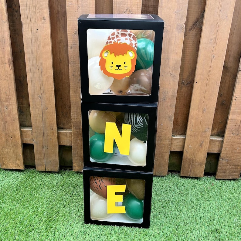 May include: Three black square boxes stacked vertically, spelling "ONE" with yellow letters. Each box contains balloons in various colours and patterns, including a lion face cutout in the top box. The boxes are set on green grass with a wooden fence background.