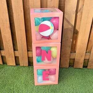 May include: Three stacked pink boxes with clear windows, filled with balloons and letters spelling "ONE". The top box features a beach ball. Balloons are pink, turquoise, and yellow. A festive decoration for a birthday celebration.
