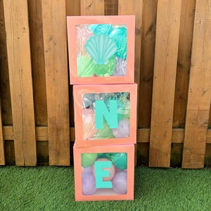 May include: Three stacked coral-colored boxes spelling "ONE" with turquoise letters. Each box has a clear window filled with balloons in shades of green, blue, and pink. The top box features a seashell cutout.