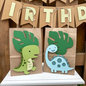 May include: Two brown paper gift bags decorated with dinosaur cutouts. One bag has a green dinosaur, the other a blue dinosaur. Each bag has a green leaf cutout and a string handle. A banner reading "BIRTHDAY" is in the background.