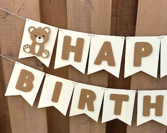 May include: A brown and white banner with the words "Happy Birthday" in brown letters. A small brown bear is on the left side of the banner.