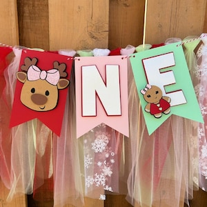 May include: A festive banner spelling "ONE" with red, pink, and green pennants. Each pennant features a letter or a holiday-themed character, including a reindeer and a gingerbread person. The banner is decorated with tulle and snowflake accents.