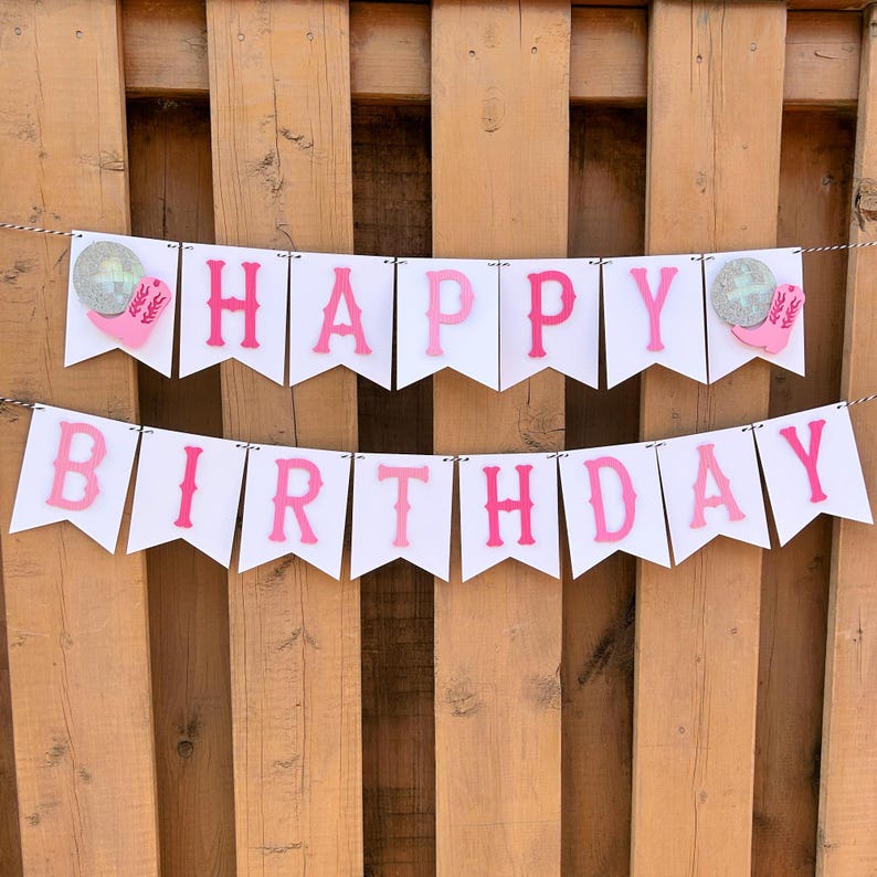 May include: A white banner with the words "HAPPY BIRTHDAY" in pink letters. The banner is strung on a thin white string and has decorative elements on each end. The background is a wooden fence.