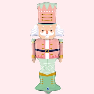 May include: A pink and green nutcracker balloon with a white beard and moustache. The balloon has a gold belt and a green hat with gold trim.