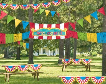 May include: A festive outdoor party scene with a red, white and blue circus-themed banner that reads "WELCOME!" hanging between trees. The banner is decorated with yellow fringe and red and white stripes. There are also colourful pennant flags and fringe garlands hanging from the trees. Two picnic tables are decorated with red, white and blue circus-themed table runners.