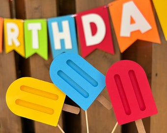 May include: Three popsicle-shaped decorations in yellow, blue, and red, each featuring two vertical recessed lines and a small wooden stick at the base. These items are positioned in front of a colorful birthday banner with white letters.