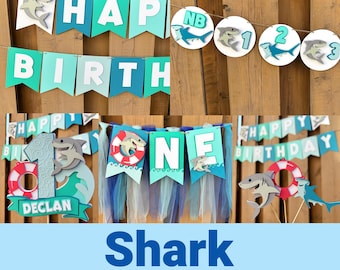 以下が含まれることがあります： 「HAPPY BIRTH」と「ONE」と書かれたバナーが付いた、サメをテーマにしたパーティーセット。装飾にはサメの切り抜き、救命浮輪、テキスト「Shark Party Bundle」が含まれています。配色は青、ティール、白です。