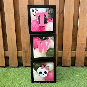 May include: Three black boxes stacked vertically, each with a clear front panel. The top box has a pink "T" and balloons. The middle box has a pink "W" and balloons. The bottom box has a skull with a pink bow and balloons.