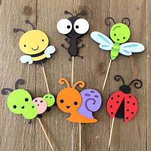 May include: A collection of colorful, cartoon-style insect cutouts on wooden sticks. The set includes a yellow bee, a black ant, a green dragonfly, a green caterpillar, an orange snail, and a red ladybug. Each insect has a smiling face.