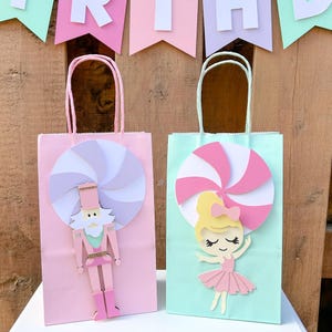 Peut inclure: Deux sacs cadeaux, un rose et un vert menthe, avec des poignées blanches. Le sac rose présente un motif de casse-noisette et une grande spirale. Le sac menthe a un motif de ballerine et une spirale. Une bannière avec le mot "BIRTHDAY" est en arrière-plan.