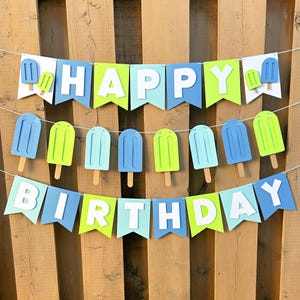 May include: A festive birthday banner with the words "HAPPY BIRTHDAY" in white letters. The banner features blue, green, and white pennants, adorned with popsicle cutouts. The banner is strung on a white string against a wooden background.