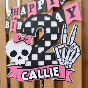 May include: A black and white checkered number two cake topper with a skull wearing a pink bow, a skeleton hand making a peace sign, and a pink banner that says "CALLIE". A "Happy Birthday" banner is in the background.