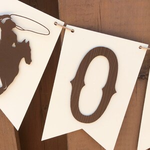 May include: A white banner with brown cut-out letters spelling "HO", a cowboy riding a horse, and a string of twine.