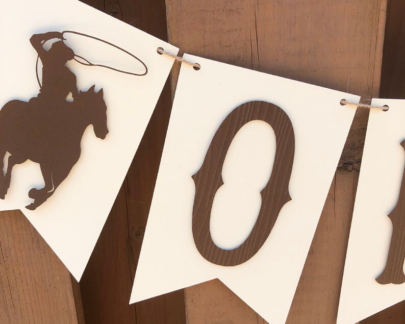 May include: A white banner with brown cut-out letters spelling "HO", a cowboy riding a horse, and a string of twine.