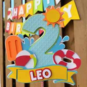 May include: A colorful birthday cake topper featuring a large number 2 in light blue with a textured pattern. The topper includes a sun, popsicle, beach ball, and life preserver. The name "LEO" is displayed on a yellow banner.