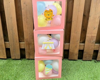Peut inclure: Trois boîtes roses empilées avec des fenêtres transparentes, chacune remplie de ballons aux couleurs pastel. La boîte supérieure présente une découpe de lion, le milieu un chapiteau de cirque et le bas une glace. Les boîtes sont sur de l'herbe verte avec une clôture en bois en arrière-plan.