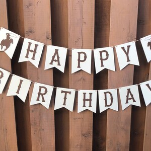 May include: White banner with brown letters spelling "Happy Birthday" with a cowboy silhouette on each end.