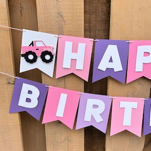 May include: A pink and purple banner with the words "HAPPY BIRTH" in white letters. The banner includes a pink monster truck graphic on a white pennant. The background is a wooden surface.
