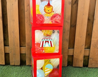 May include: A stack of three square red display boxes with clear windows, each filled with colourful balloons. The top box features a lion graphic, the middle box shows a circus tent, and the bottom box displays an ice cream cone graphic.