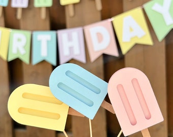 May include: Three popsicle-shaped cutouts in pastel yellow, light blue, and soft pink, each featuring two vertical indentations and a wooden stick. In the background, a pennant banner displays the letters of the word BIRTHDAY.