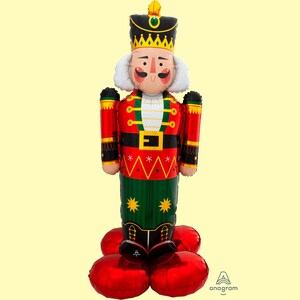 May include: A red and green inflatable nutcracker balloon with a black hat and a gold crown. The balloon has a white collar and a black belt with a gold buckle. The balloon is standing on two red inflatable balloons.