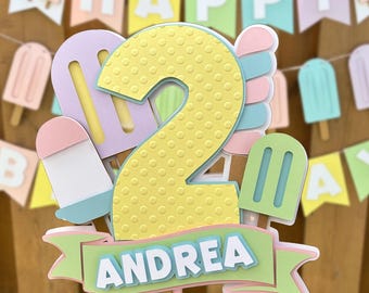 May include: A cake topper featuring a large yellow number two with a dotted texture, surrounded by pastel-colored popsicle cutouts. A light green banner at the bottom displays the name ANDREA in white capital letters.