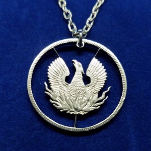 Puede incluir: Un collar de plata con un colgante de moneda que presenta un recorte detallado de un fénix que surge de las llamas.