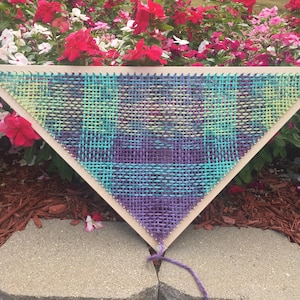 May include: A triangular loom with a woven tapestry in shades of purple, blue, and green. The loom is made of wood and has a string attached to the bottom.