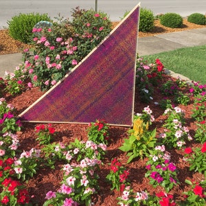 May include: A triangular shaped metal sculpture with a purple, gold, and blue iridescent finish. The sculpture is surrounded by a bed of pink, white, and red flowers.