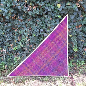 May include: A triangular knitted shawl in shades of purple, pink, and gold. The shawl is laid out on a bed of green ivy.