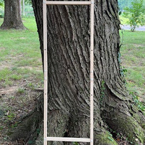 May include: A wooden frame with three horizontal bars, leaning against a tree trunk. The frame is made of natural wood and has a rustic appearance.