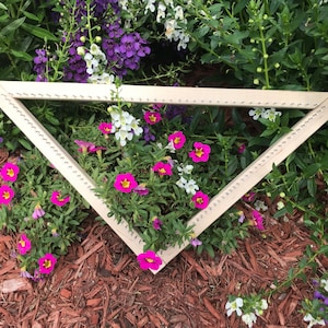 May include: A wooden triangular loom with a row of nails along each side, surrounded by pink, white and purple flowers.