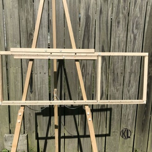 May include: A wooden loom with two frames, set up on a wooden easel. The loom is made of natural wood and has a simple design.