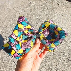 May include: A large, colorful hair bow featuring a patchwork design in shades of yellow, teal, pink, and black. The bow is held together with a black band. The bow is made of a patterned fabric.