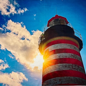 May include: A tall lighthouse with red and white horizontal stripes, topped with a red roof and a black band. The sun shines brightly behind the lighthouse, with a blue sky and fluffy white clouds in the background.