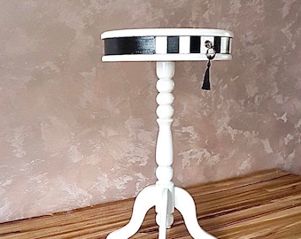 Puede incluir: Mesa auxiliar blanca con una tapa a cuadros en blanco y negro y un borla. La mesa tiene una tapa redonda y tres patas.