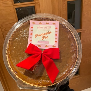 May include: A round pumpkin pie in a clear plastic container, topped with a bright red fabric bow. A square label features the text The Happy Bakery Co. Pumpkin Pie and 650g, surrounded by small orange pumpkin icons on a light pink background.
