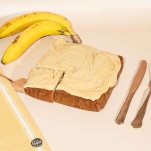 May include: A square piece of banana bread topped with a thick layer of vanilla frosting. Two ripe bananas and a butter knife are on the table next to the bread. Two gold forks are on the table to the right of the bread.